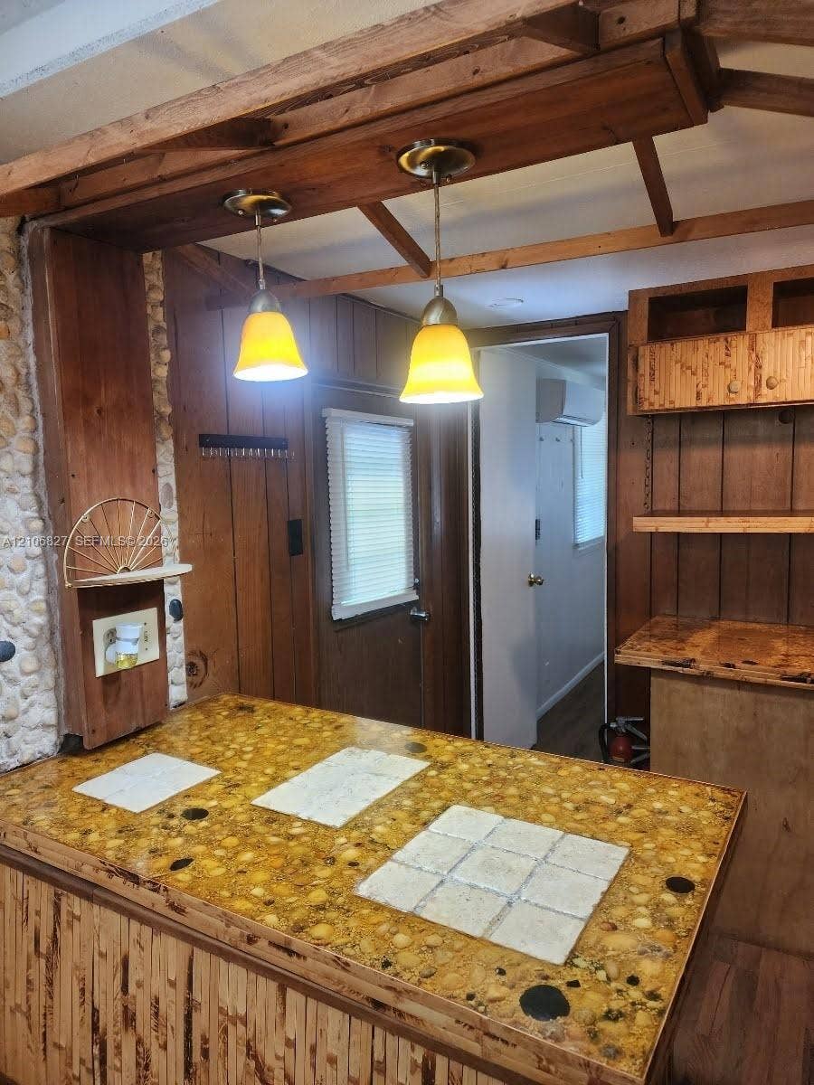 Kitchen island featuring a textured stone countertop with integrated tile insets, wood-paneled base cabinetry, dual pendant lighting, and overhead wooden beam detailing