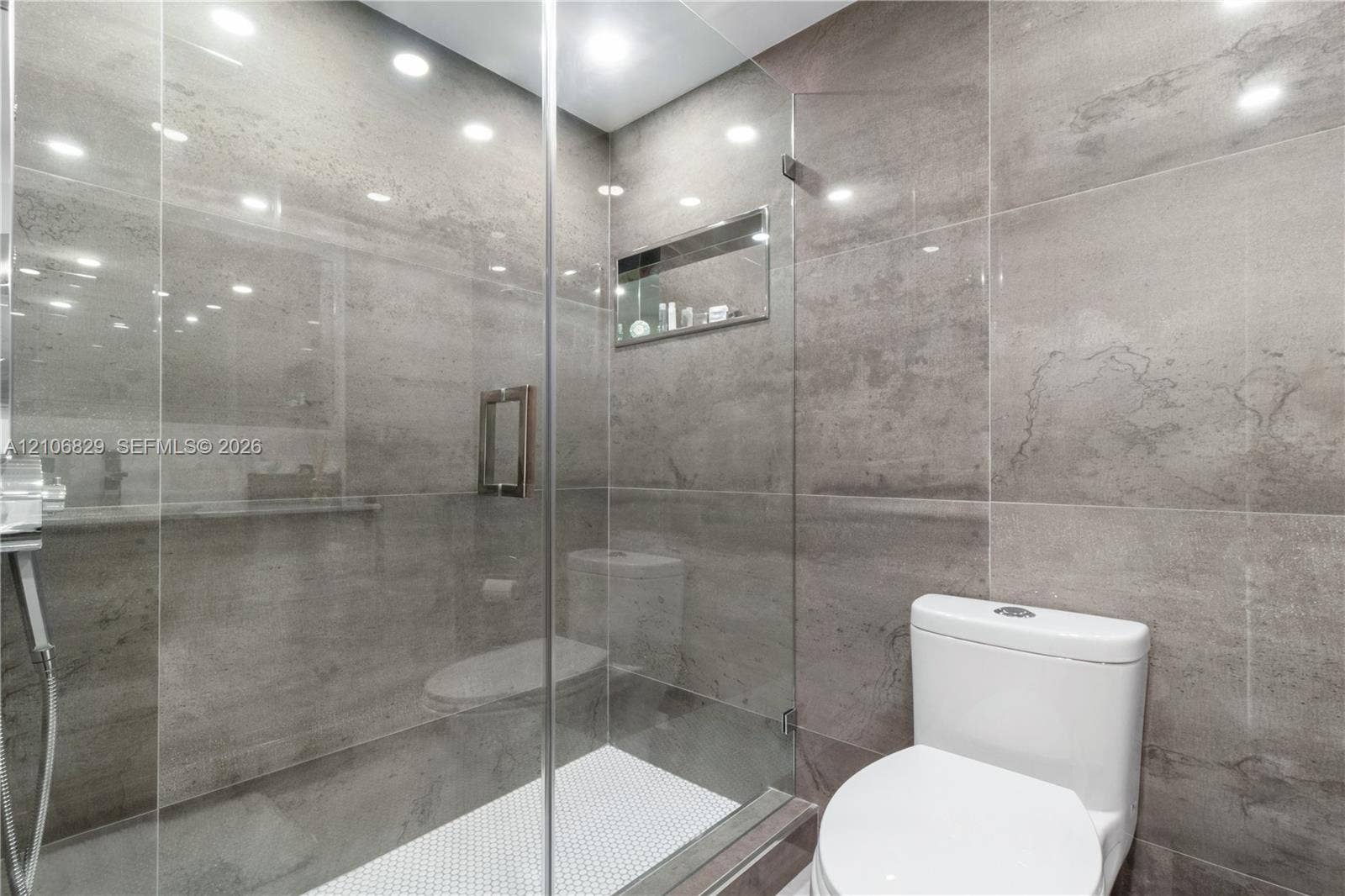 Walk-in shower featuring large-format grey tile, a frameless glass enclosure, a built-in recessed shelf, and white mosaic floor tile, accompanied by a dual-flush toilet