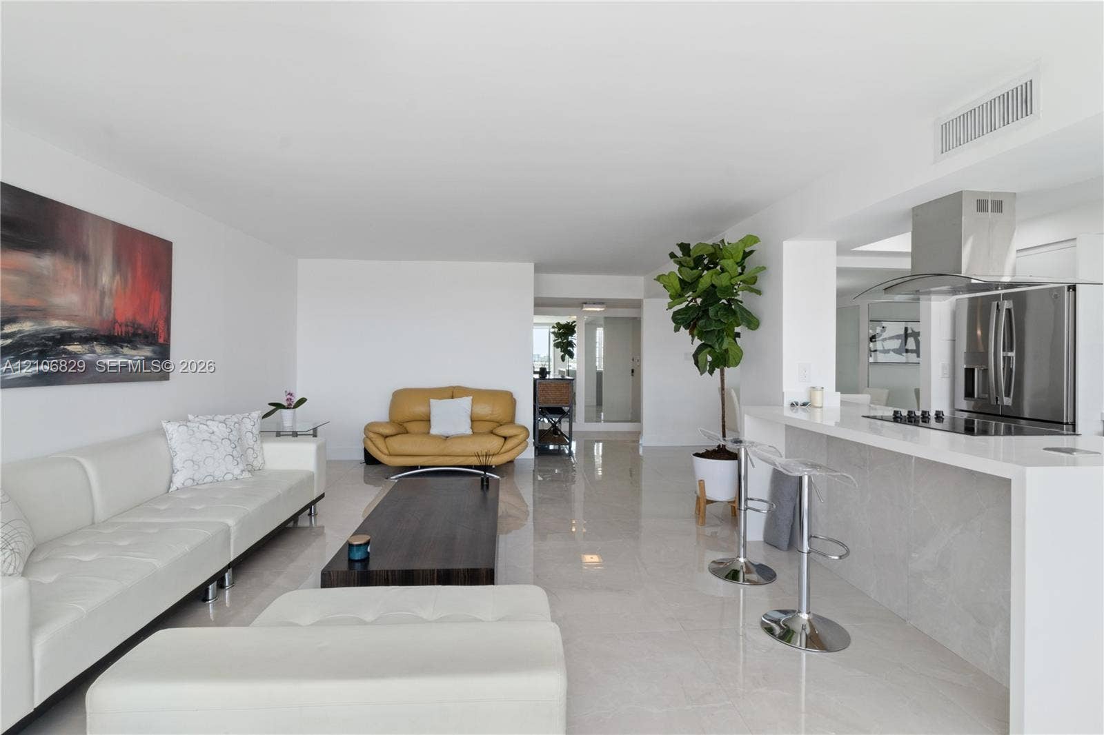 Open-concept interior featuring light-toned tile flooring, waterfall-edge countertop, stainless steel refrigerator, and built-in stainless steel range hood