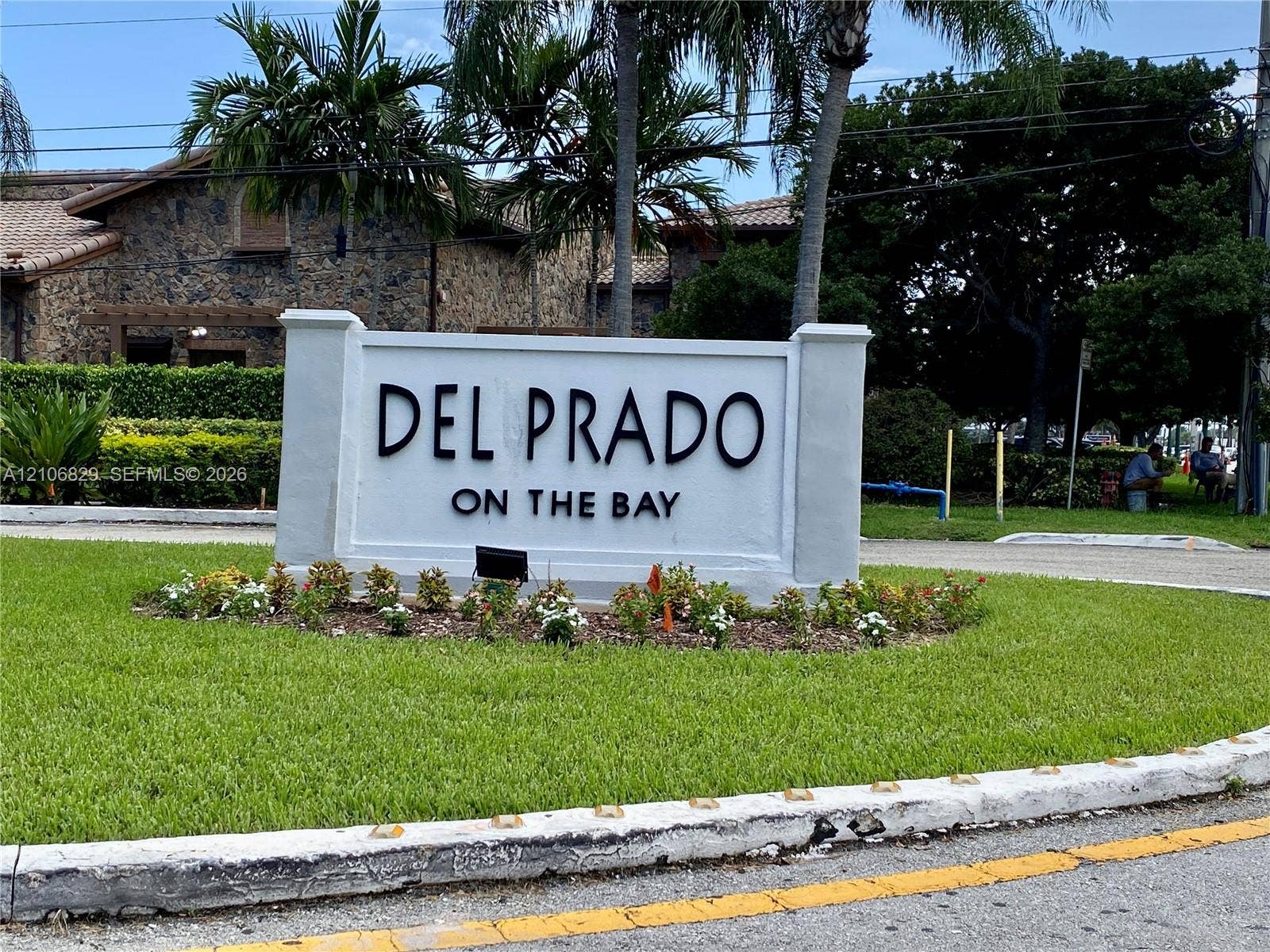 Community entrance signage featuring raised dimensional lettering, surrounded by manicured landscaping, mature palms, and structured masonry columns