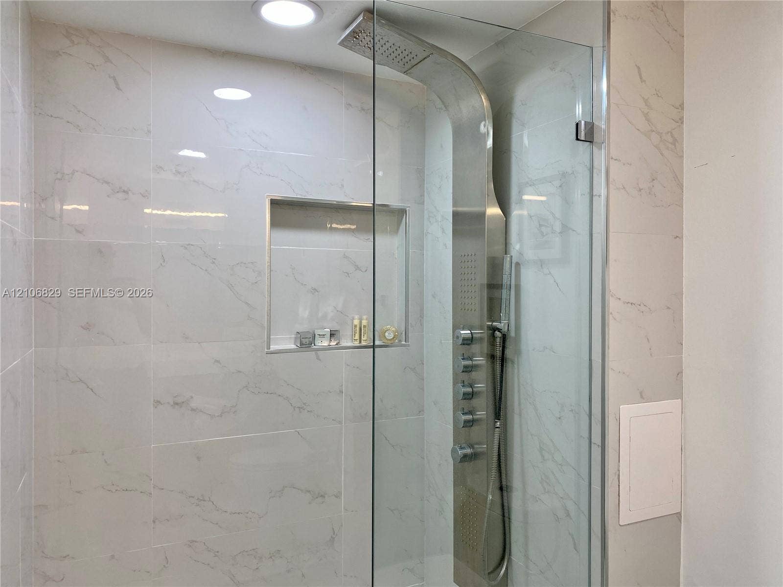 Walk-in shower featuring large-format stone-finish tile surround, integrated shower panel system, rain shower head, handheld spray, and a recessed wall niche with glass door enclosure