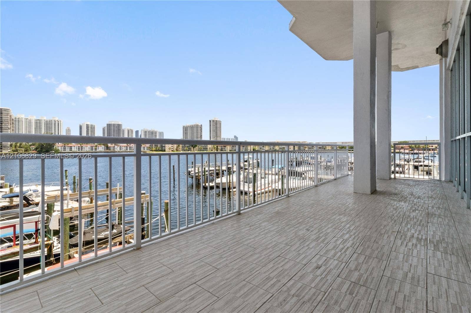 Covered balcony featuring wood-finish tile flooring, white structural pillars, and a metal railing overlooking a waterway with boat slips
