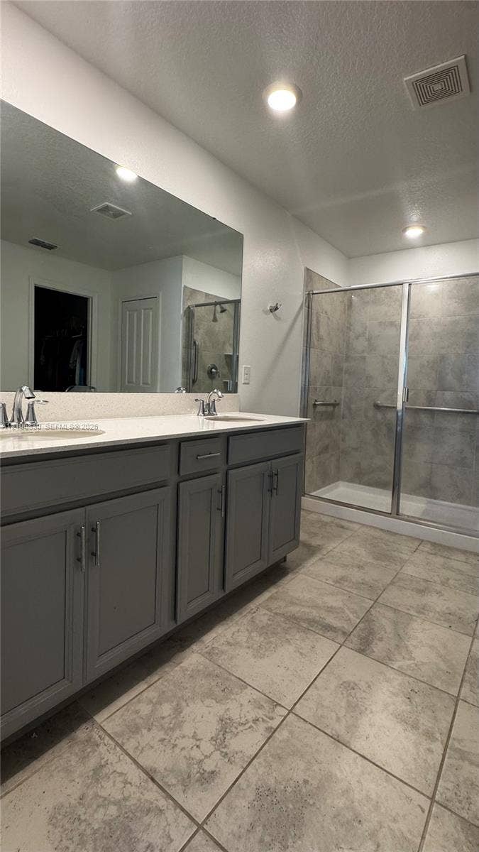 Dual-sink vanity with gray shaker-style cabinetry, white countertop, large wall-to-wall mirror, and glass-enclosed shower with stone-finish tile surround