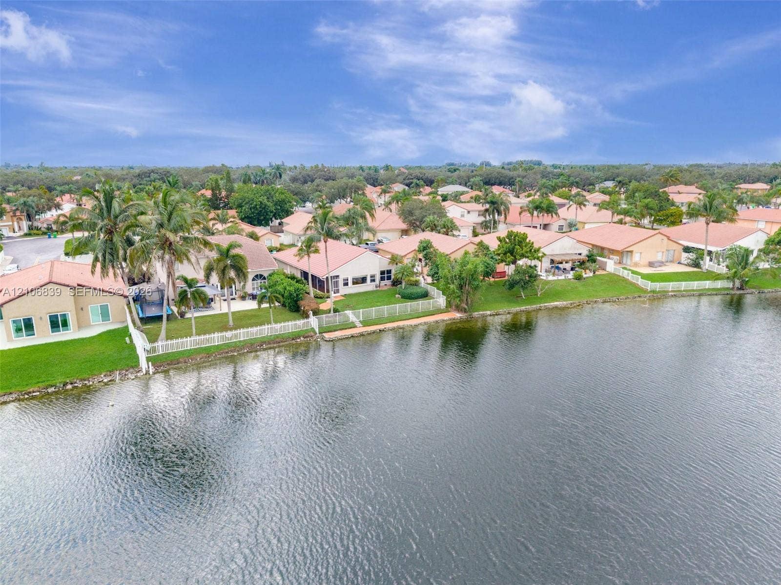 12683 NW 11th Place, Sunrise, FL 33323 Waterfront property featuring a private white picket fence, mature palm trees, manicured lawns, and concrete patio surfaces
