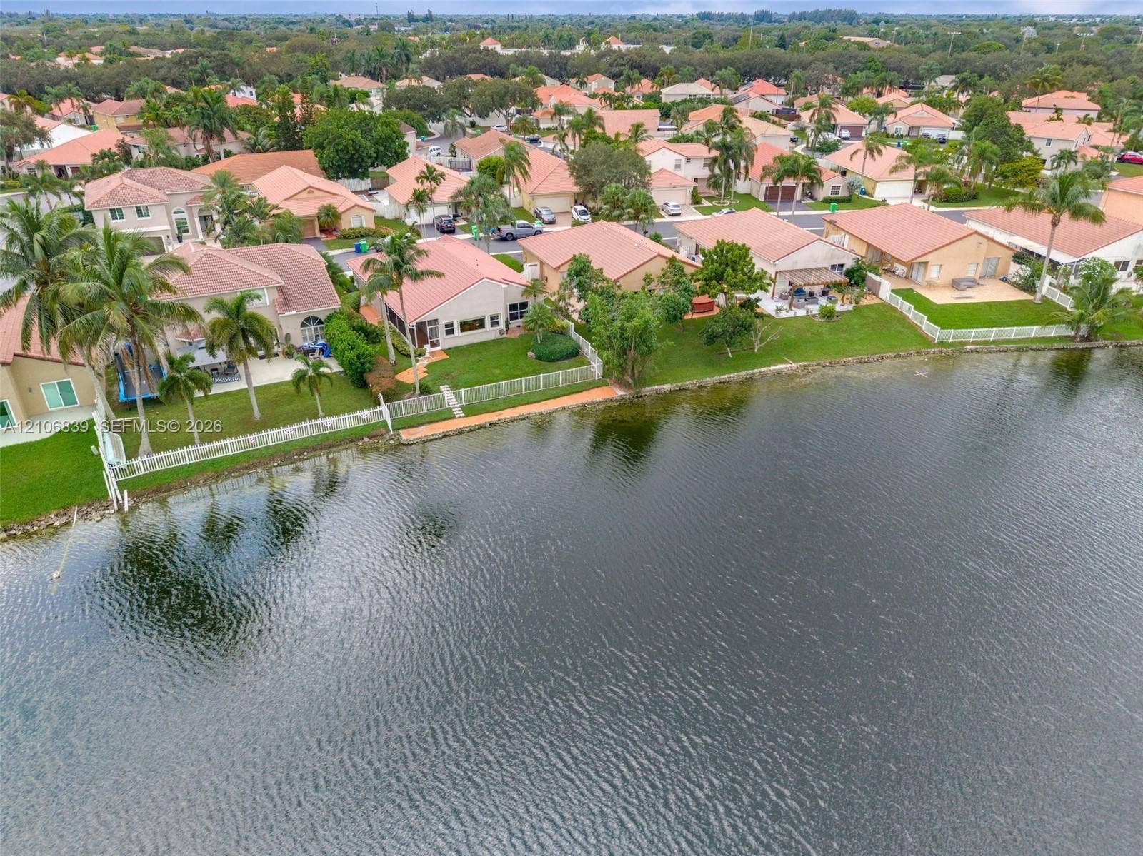 12683 NW 11th Place, Sunrise, FL 33323 Waterfront residential setting featuring a large body of water, manicured lawns, white vinyl fencing, and palm trees