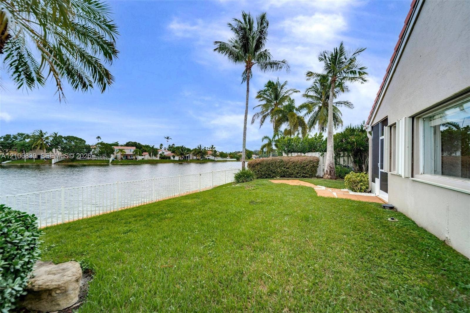 12683 NW 11th Place, Sunrise, FL 33323 Waterfront exterior featuring a grass lawn, metal fencing, stone-finish paving, and mature palm trees