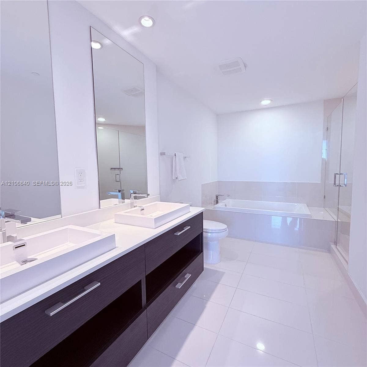 Dual vanity with rectangular vessel sinks and stone-composite countertop, dark wood-finish cabinetry, recessed ceiling lighting, tiled soaking tub, and glass-enclosed shower