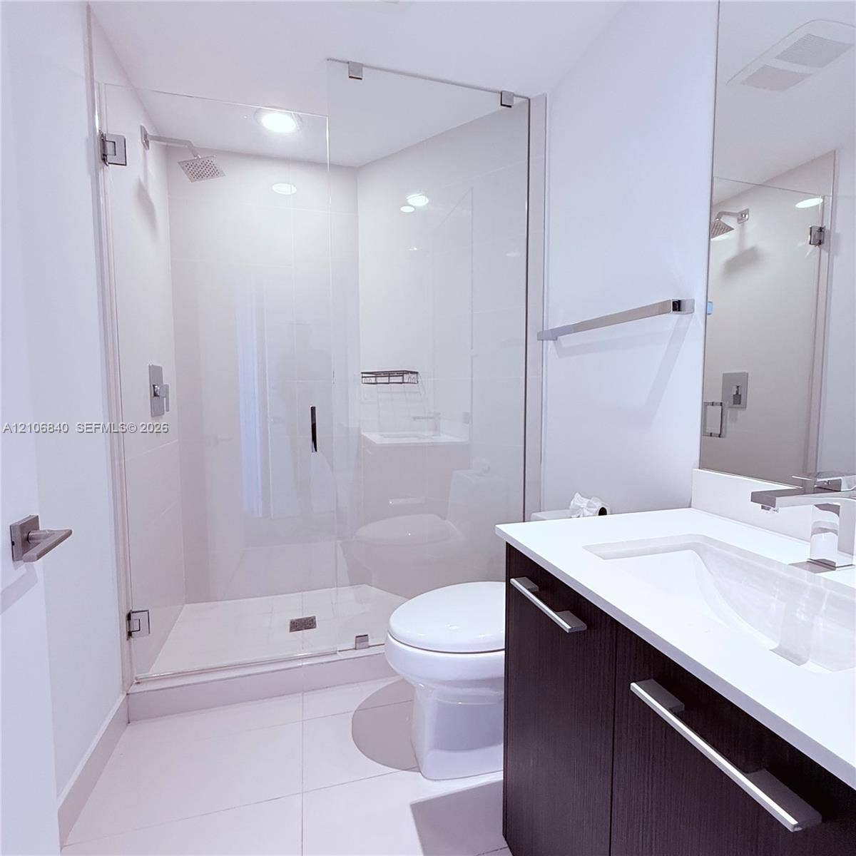 Walk-in shower with frameless glass enclosure, dark-finish cabinetry, solid-surface vanity countertop with integrated sink, polished chrome faucet, and rectangular floor tiling