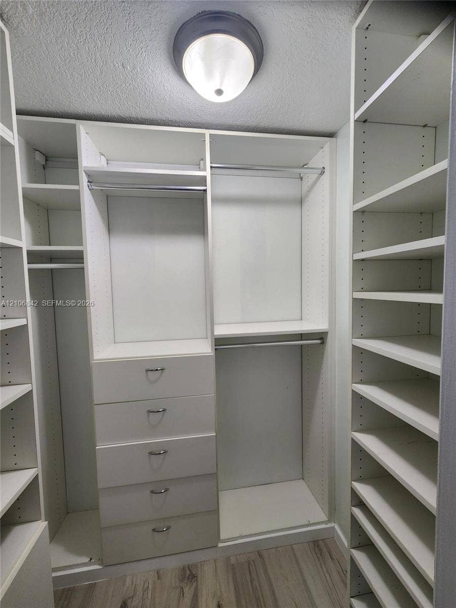 Customized storage closet featuring white built-in shelving, integrated hanging rods, a five-drawer dresser unit, and wood-finish flooring