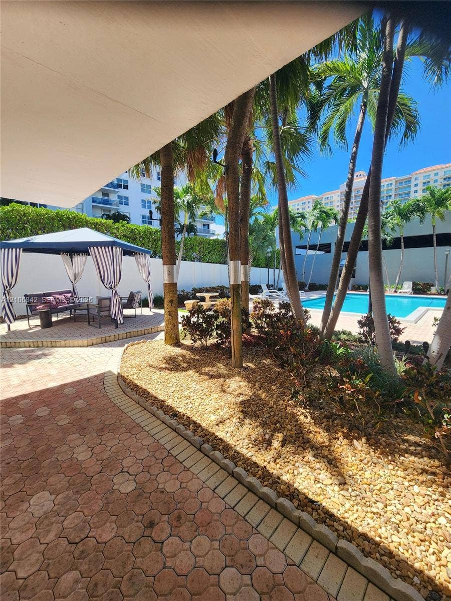 Outdoor patio area featuring interlocking paver hardscaping, a metal-frame gazebo, a swimming pool, and mature palm trees
