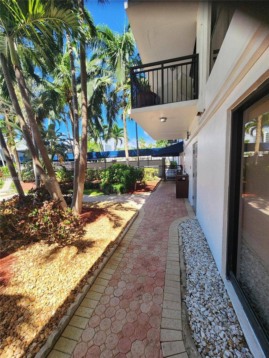 Exterior walkway featuring interlocking stone pavers, a private balcony with black metal railings, and established tropical landscaping