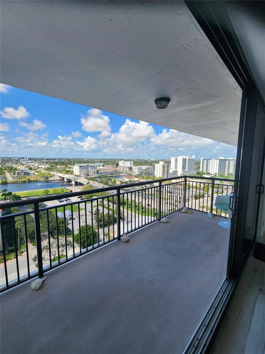Covered balcony with black metal railings and concrete flooring, featuring an expansive exterior outlook over the surrounding area, water, and cityscape