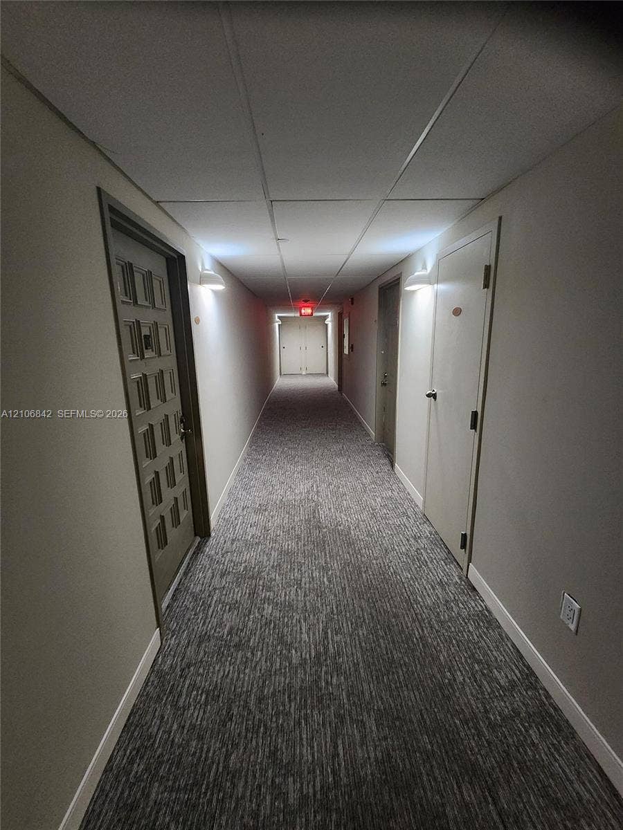 Interior hallway featuring neutral walls, grey patterned carpeting, textured drop ceiling tiles, and wall-mounted sconce lighting