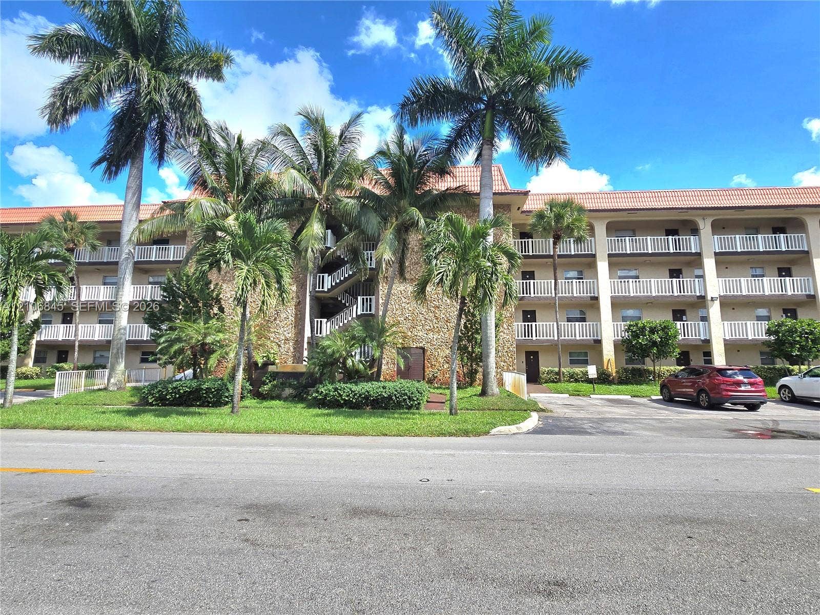 1700 NW 58th Terrace, #3J, Sunrise, Sunrise, FL 33313 Multi-story building exterior featuring stone masonry cladding, a terracotta tile roof, and wrap-around balconies with white railings, set amidst mature palm trees and landscaped grounds