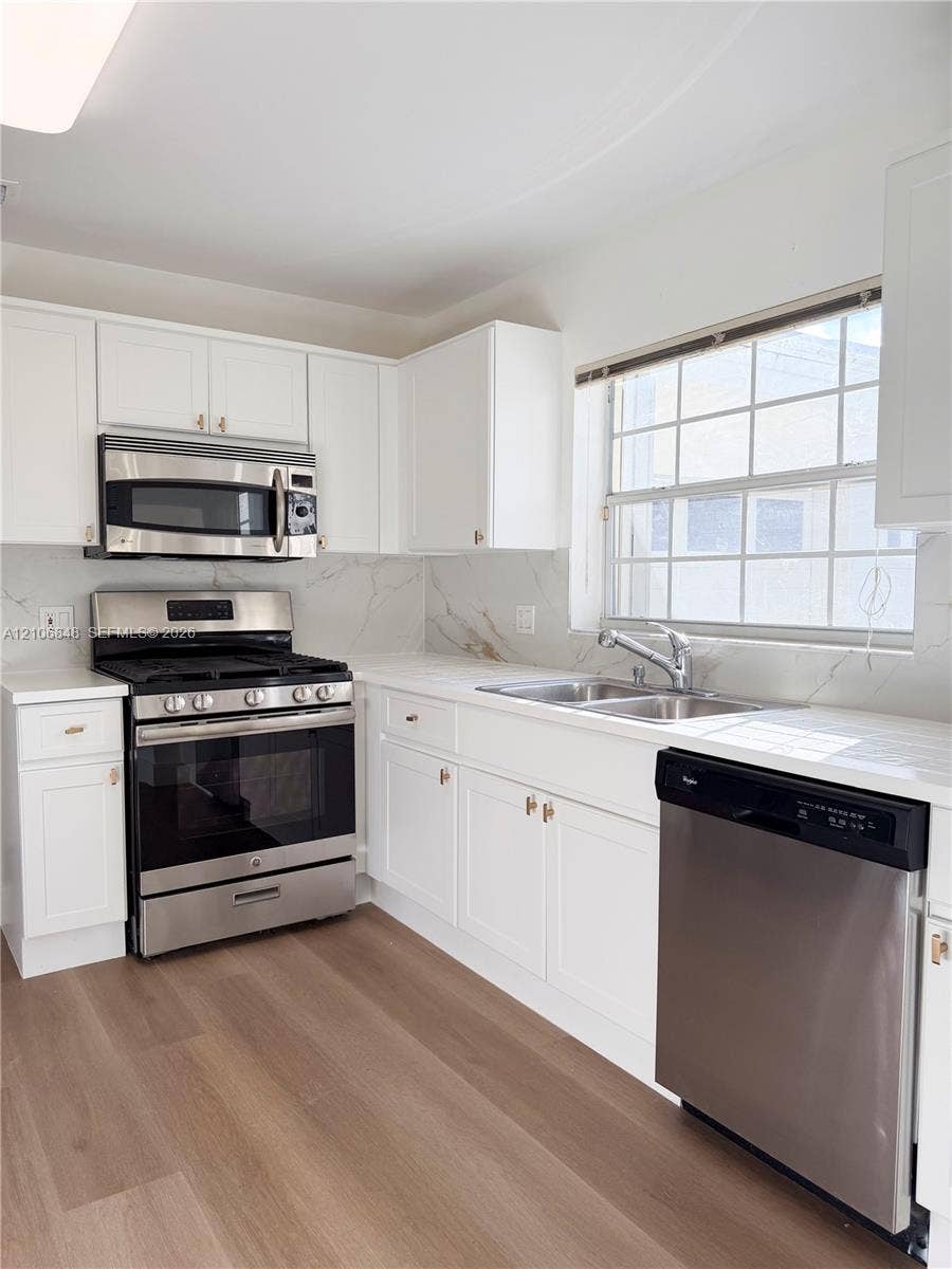 Kitchen featuring white shaker-style cabinetry, stone-surface countertops, full-height stone backsplash, stainless steel appliances, and wood-finish flooring