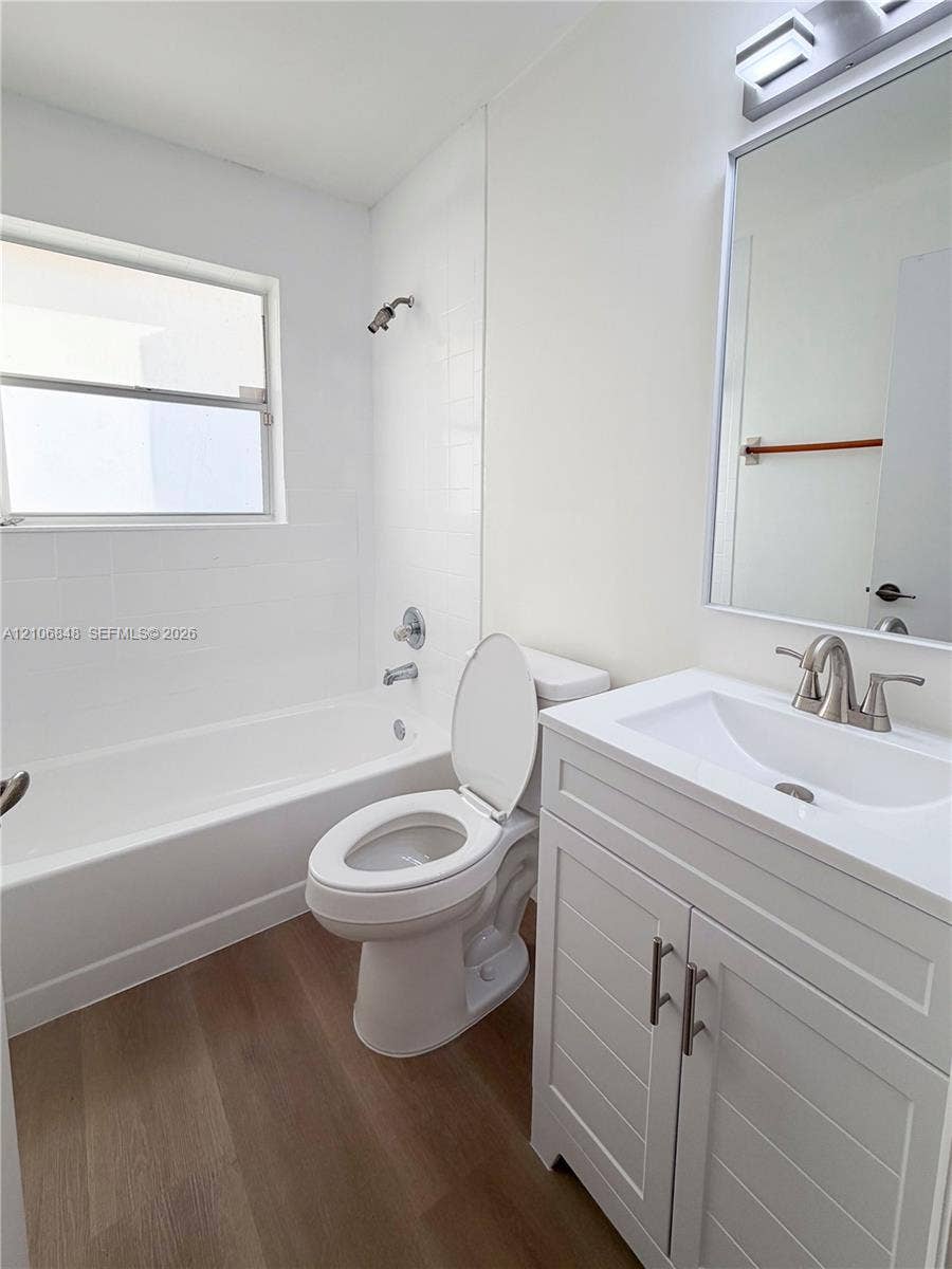 Bathroom featuring wood-finish flooring, white cabinetry with silver-tone hardware, a white-tiled shower surround, a rectangular wall-mounted mirror, and a single-basin sink with a metallic faucet
