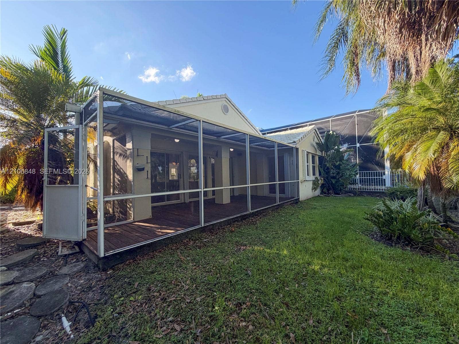 Screened-in rear porch featuring wood-finish decking and a metal frame, set against a manicured lawn with mature palm trees and established landscaping