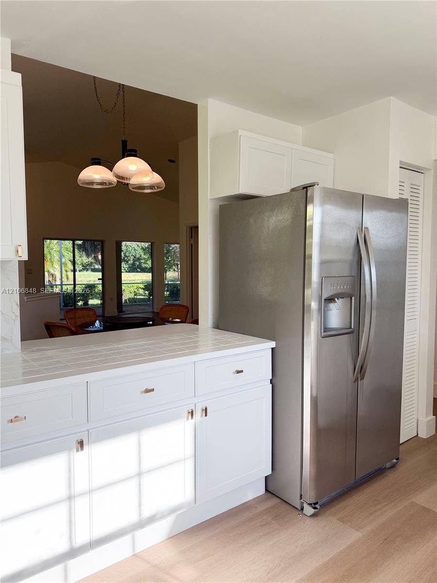 Kitchen featuring white Shaker-style cabinetry, tile countertops, stainless steel side-by-side refrigerator, multi-light pendant fixture, and light-toned wood-finish flooring