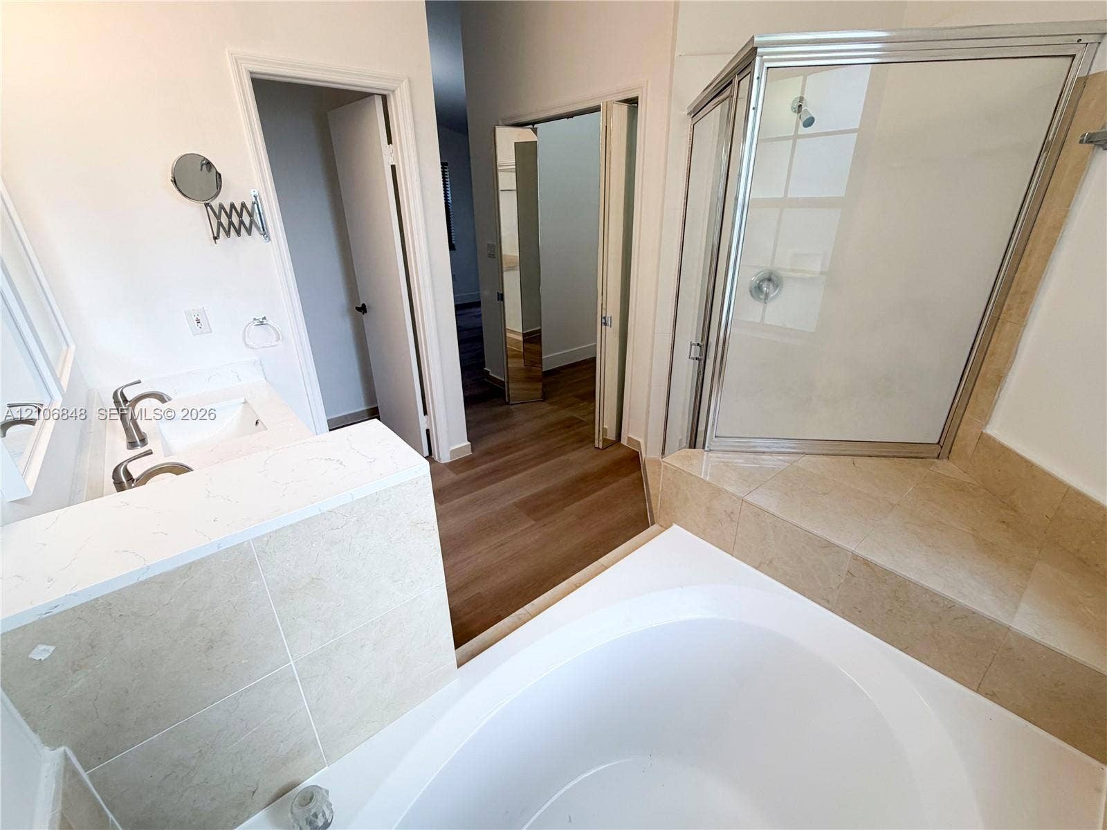 Bathroom featuring a quartz-topped vanity, white soaking tub, corner glass-enclosed shower, stone-tile surrounds, and wood-finish flooring