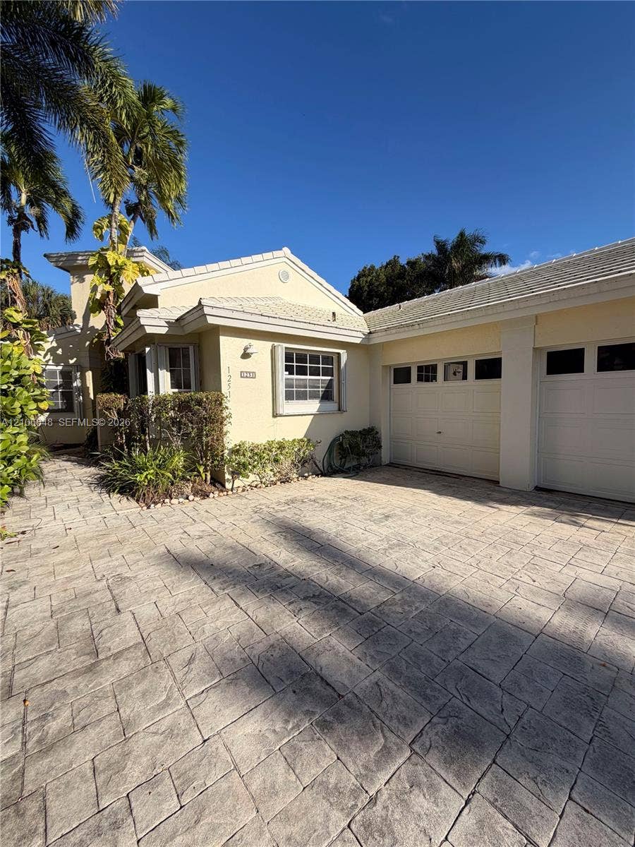 Stucco exterior featuring a dual-car garage, patterned stone paver driveway, tiled roof, and mature palm landscaping