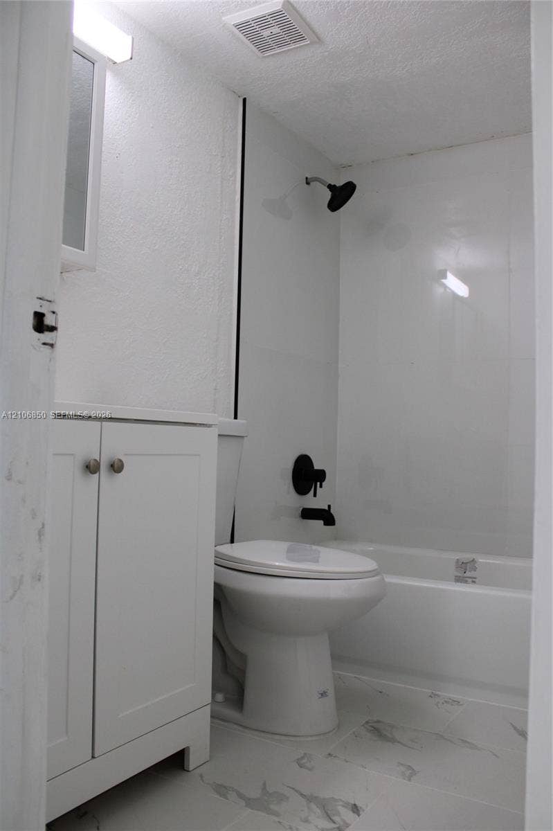 Bathroom featuring a white vanity cabinet, bathtub with a shower enclosure, and stone-finish tile flooring complemented by matte black plumbing fixtures