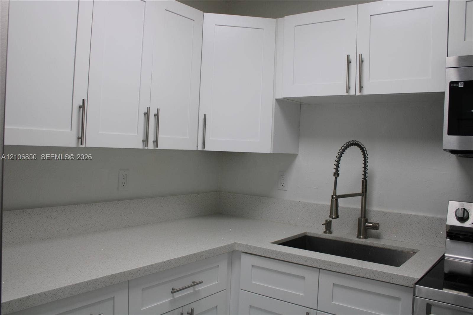 White Shaker-style cabinetry with polished metal hardware, light-toned solid surface countertops, a stainless steel undermount sink, and a high-arch pull-down faucet