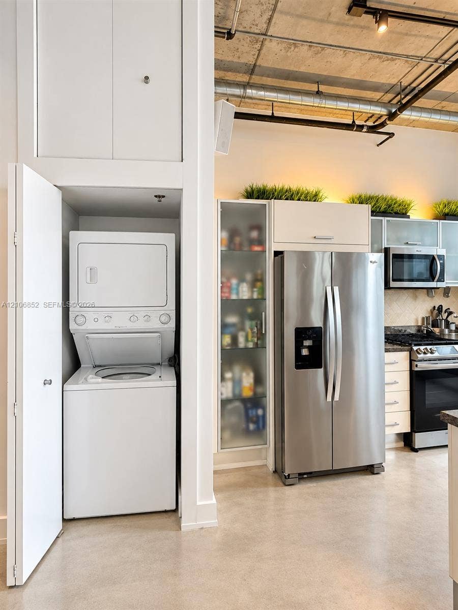 Exposed concrete ceiling, stacked laundry unit with overhead cabinetry, stainless steel appliances, glass-front cabinetry, and polished concrete flooring