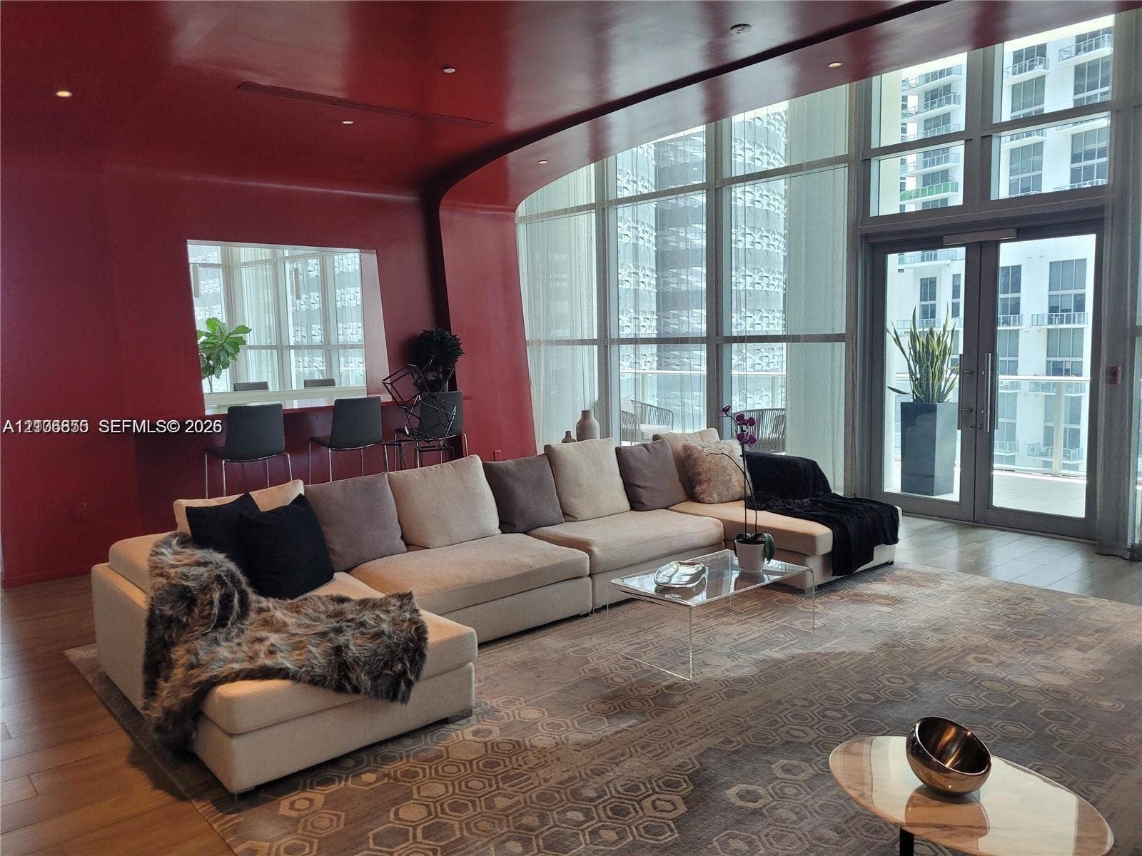 Floor-to-ceiling glass walls, integrated bar with counter seating, high-gloss red accent wall, recessed ceiling lighting, and wood-finish flooring