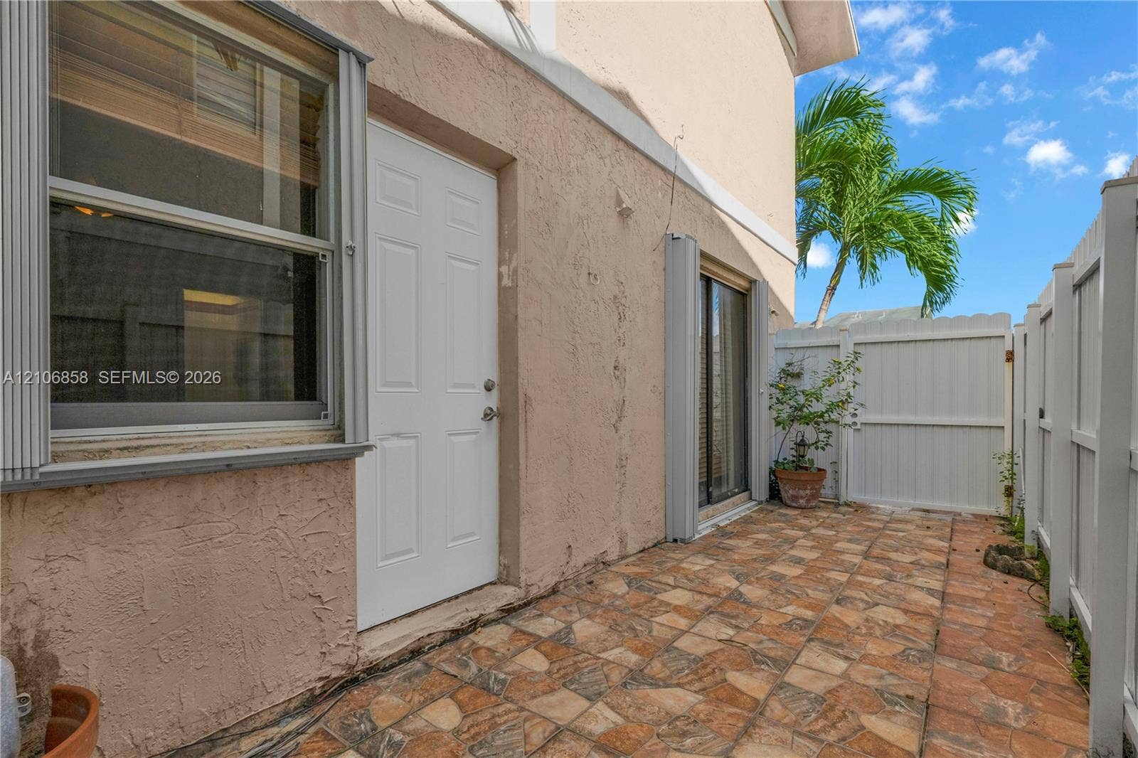 10960 SW 25th Street, Miami, FL 33165 Stucco exterior with multi-pane window, white paneled entry door, sliding glass door, and tiled patio enclosed by white vinyl fencing