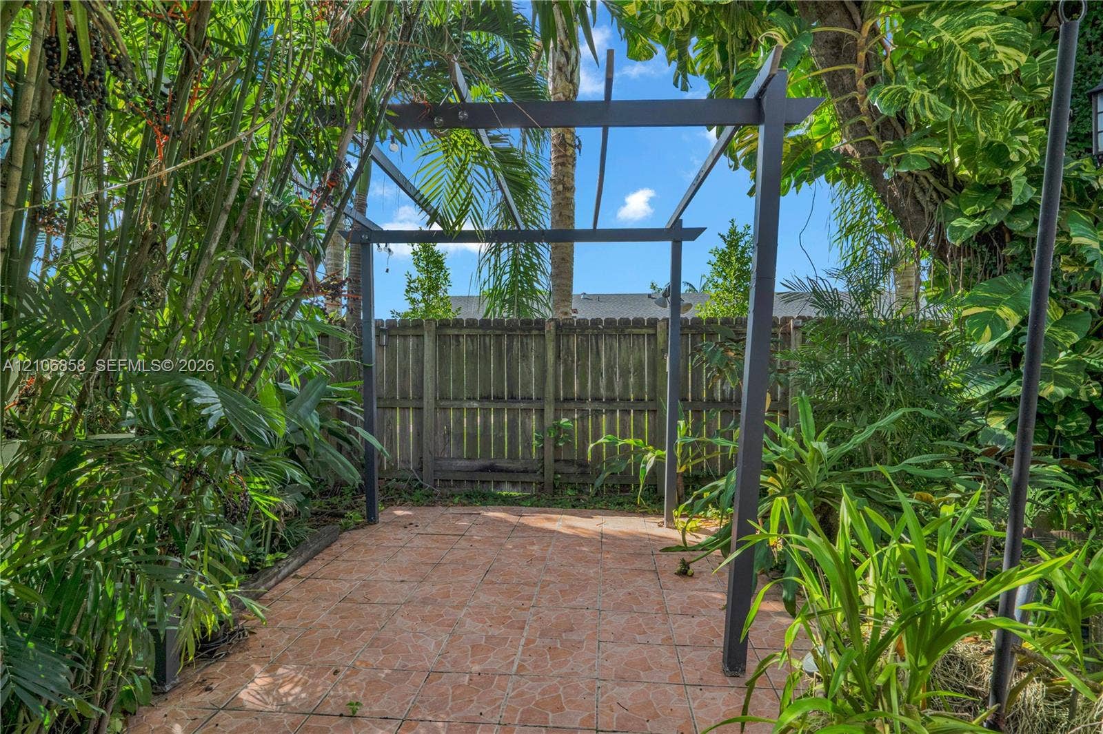 10960 SW 25th Street, Miami, FL 33165 Outdoor patio area featuring a stone-tile floor, a dark-finished metal pergola, and perimeter wood fencing surrounded by dense mature vegetation