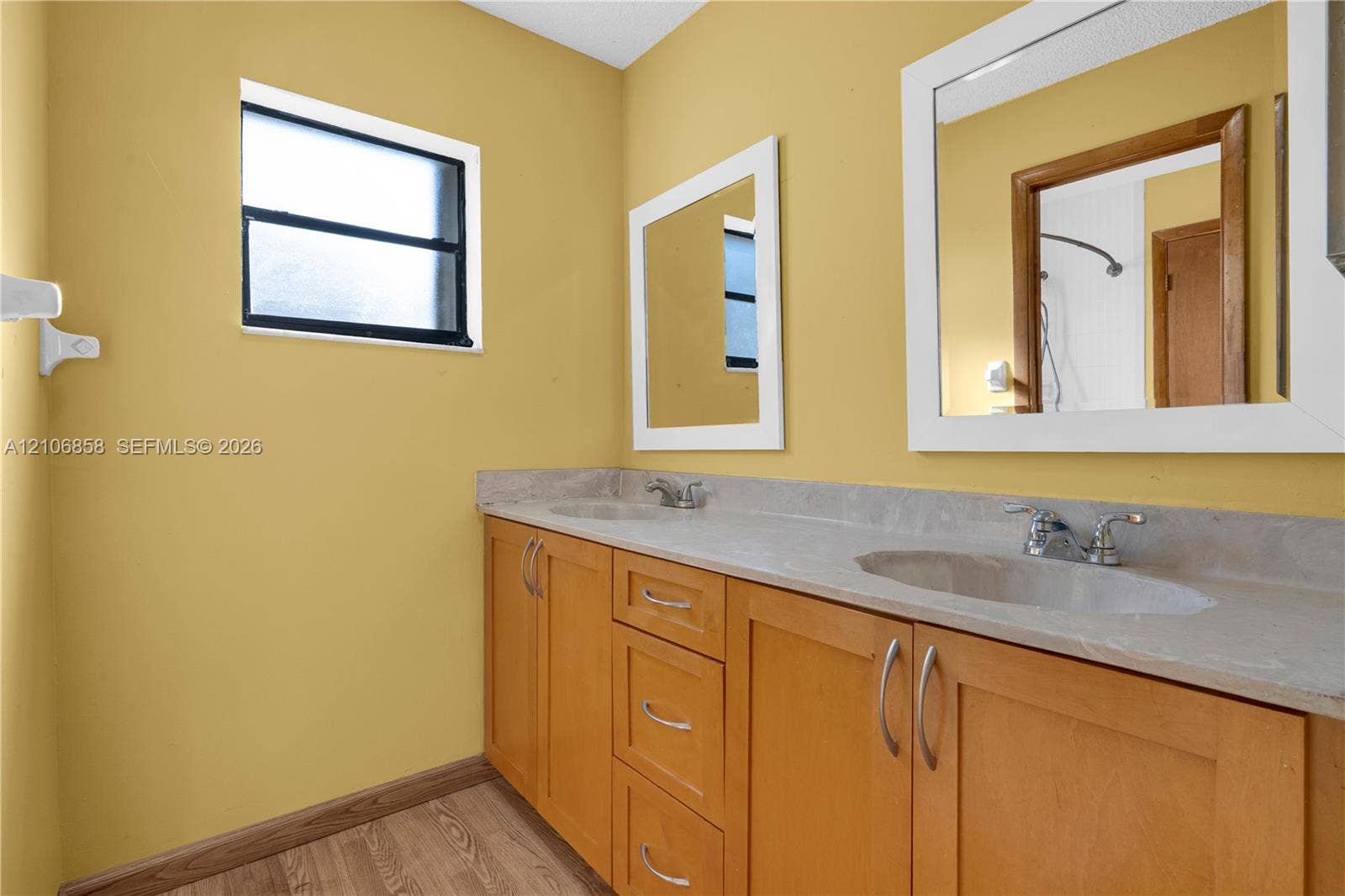 10960 SW 25th Street, Miami, FL 33165 Dual-sink vanity with a solid-surface countertop, wood-finish cabinetry, rectangular framed mirrors, and a window providing natural light