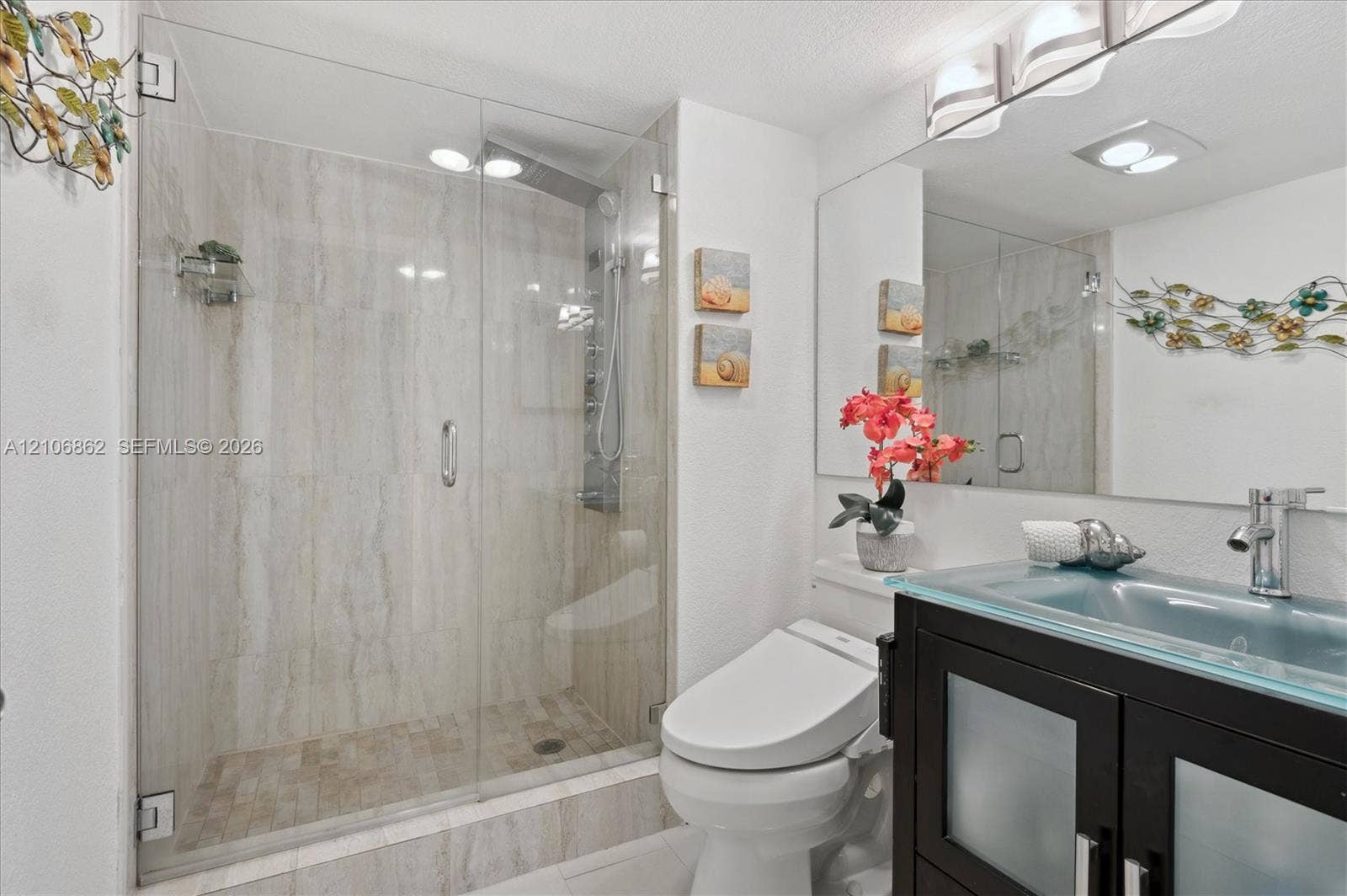 Walk-in shower with floor-to-ceiling stone-look tiling and frameless glass door, vanity with glass countertop and integrated sink, and dark wood-finish cabinetry with frosted glass inserts
