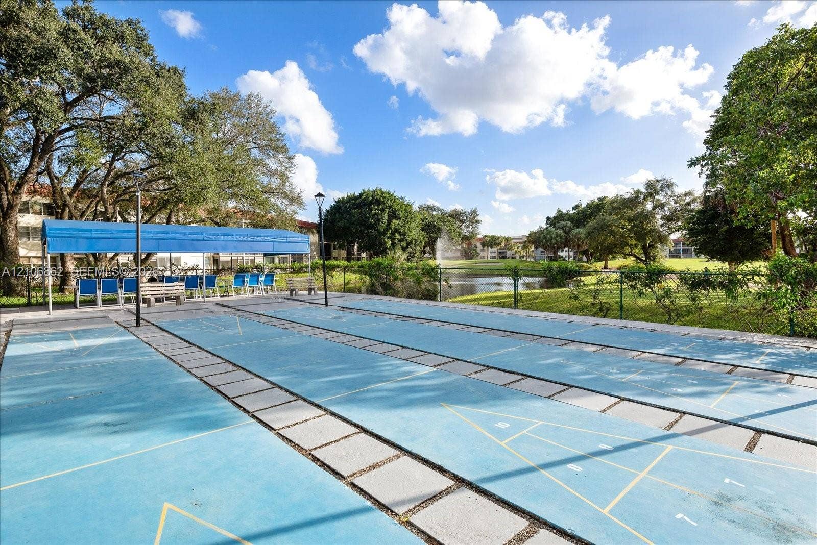 Outdoor shuffleboard courts featuring a paved surface, blue canopy shading, built-in light posts, and proximity to a water feature
