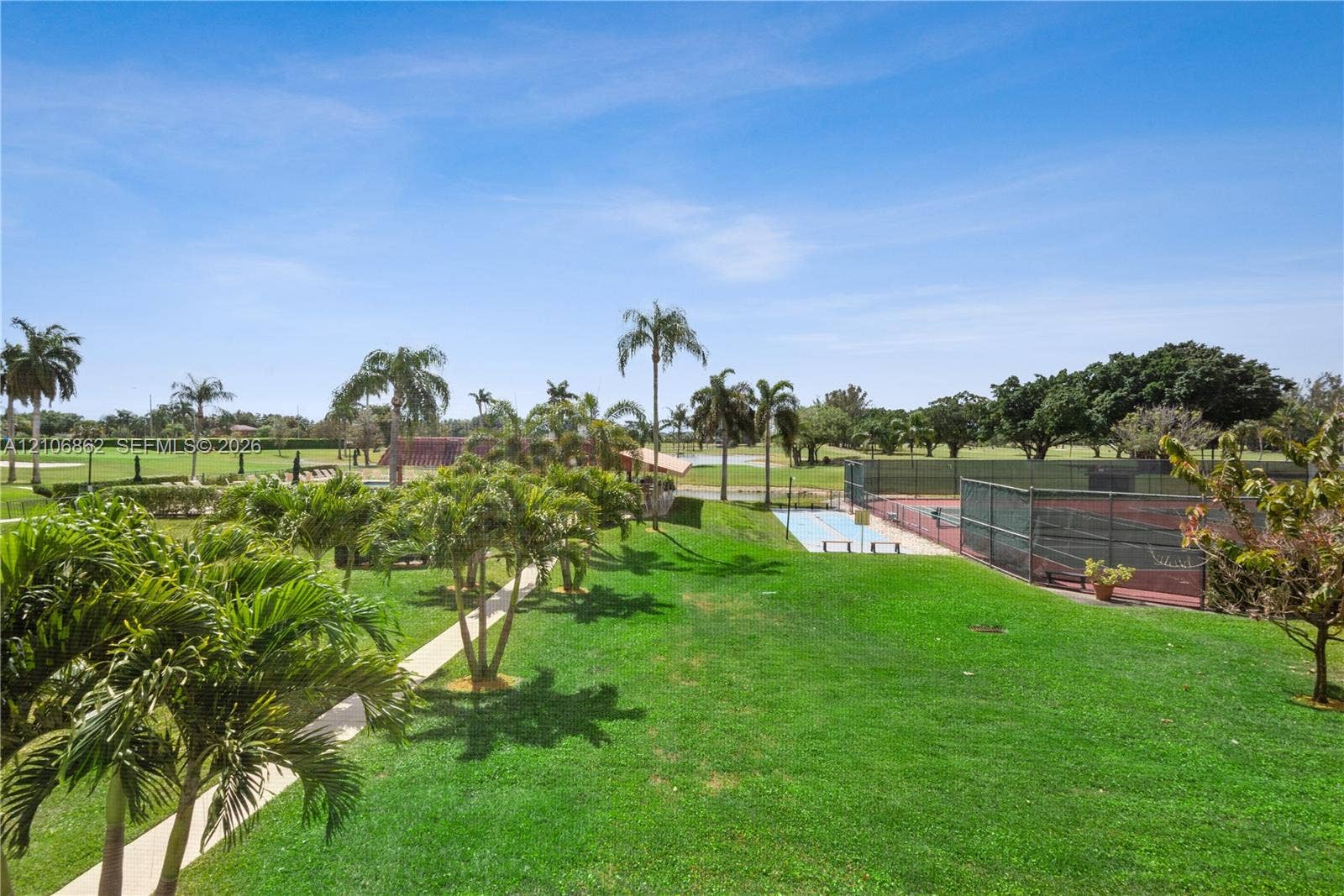 Expansive grounds featuring a fenced tennis court, manicured lawn, concrete walkways, and mature palm trees