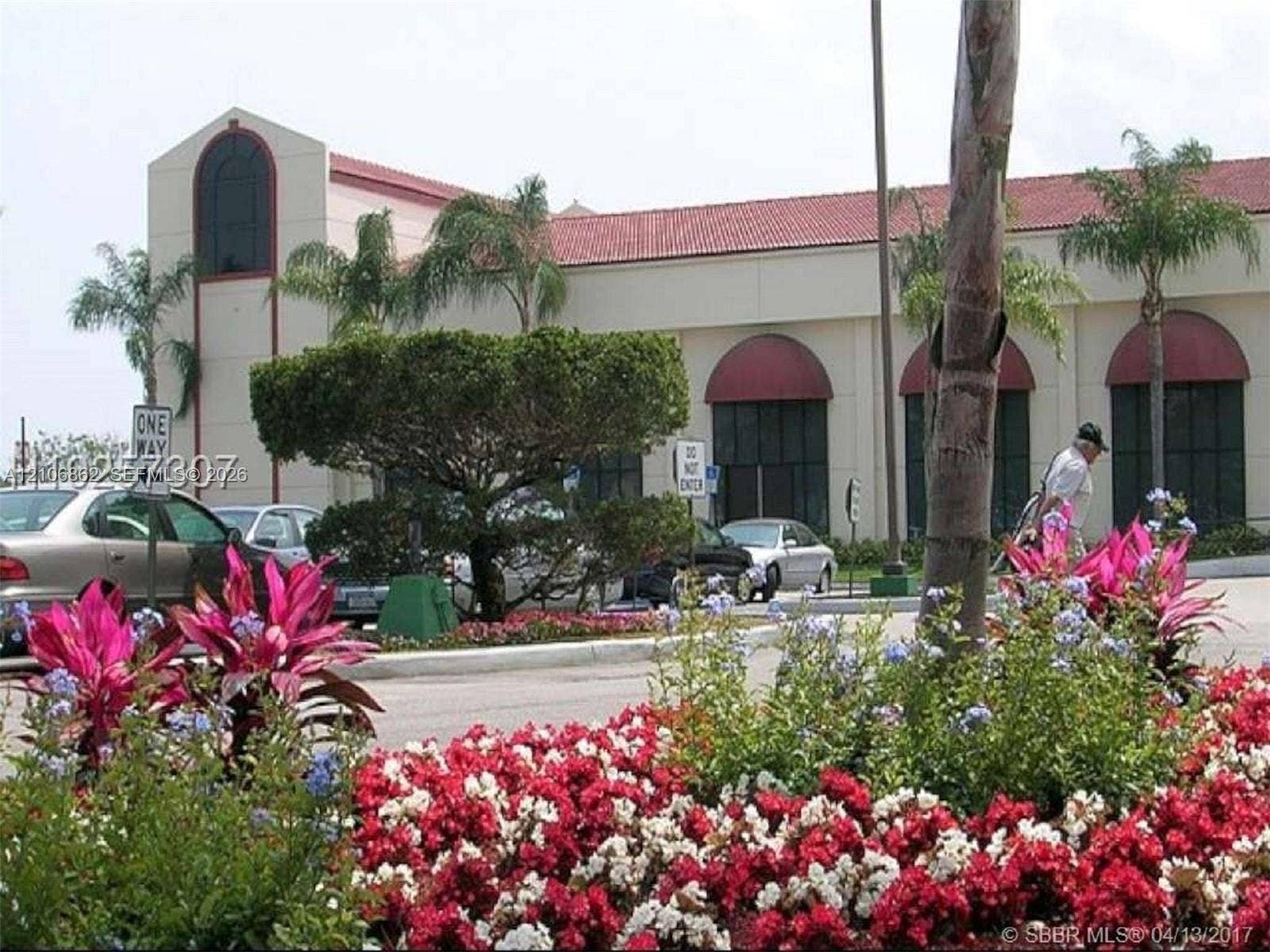 Commercial exterior featuring a light-toned stucco faade, red clay tile roof, arched window treatments, and landscaped grounds with mature palm trees