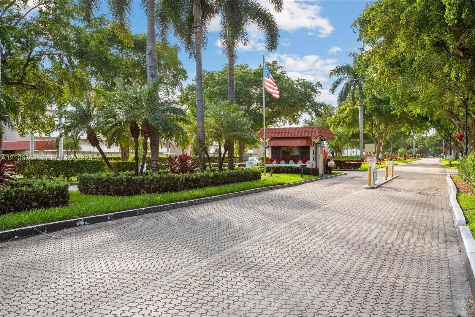 Paved entrance featuring a controlled-access gatehouse, mature palm trees, manicured hedges, and professional landscaping
