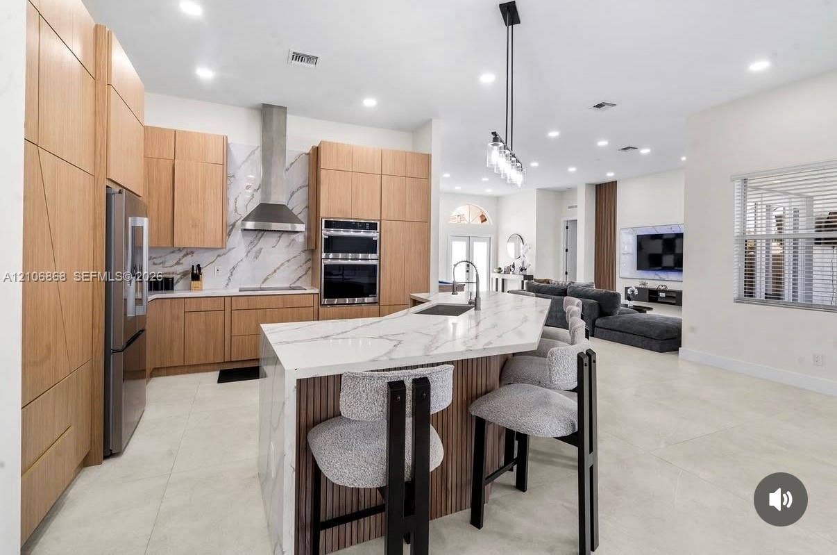 Kitchen featuring flat-panel wood-finish cabinetry, stone-finish waterfall edge island, stainless steel wall-mounted oven and microwave, stone-finish backsplash, and recessed ceiling lighting