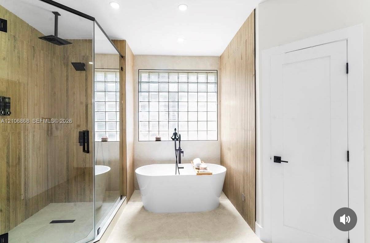 Glass-enclosed shower with matte black rainfall showerhead, freestanding soaking tub with floor-mounted faucet, wood-finish wall paneling, and glass block window