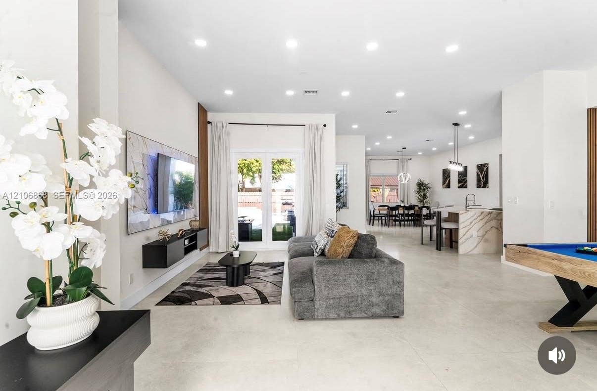Open-concept interior featuring large-format tile flooring, recessed ceiling lighting, a wall-mounted floating media console, and a kitchen island with a waterfall-edge countertop
