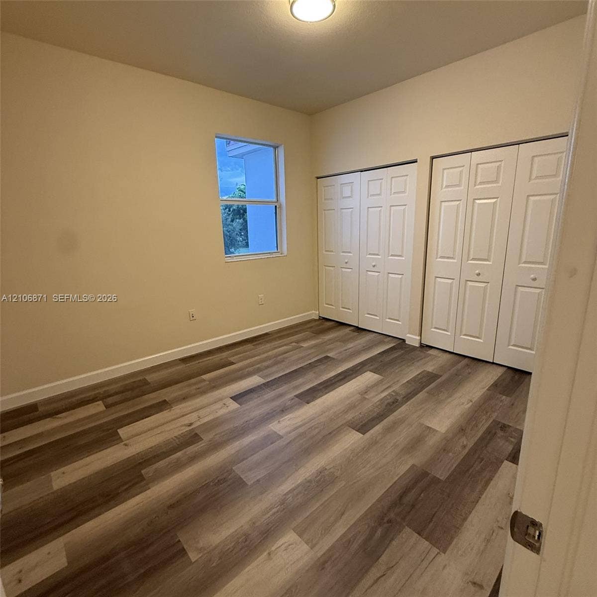 Room featuring wood-finish flooring, dual bi-fold door closets, a single-pane window, and a flush-mount ceiling light fixture