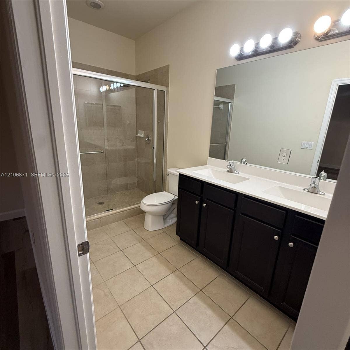 Bathroom featuring a double-sink vanity with dark-finish cabinetry, a solid-surface countertop, a large wall-mounted mirror, and a walk-in glass-enclosed shower with tile surround