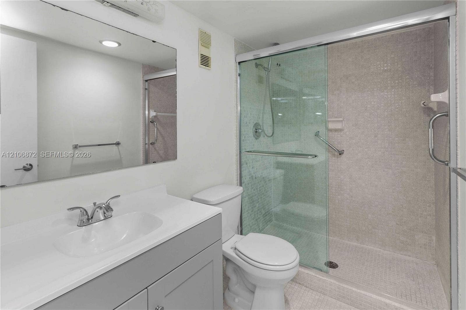 Bathroom featuring a vanity with a solid-surface countertop, an oversized wall mirror, a walk-in shower with glass sliding doors, and mosaic tile shower wall cladding