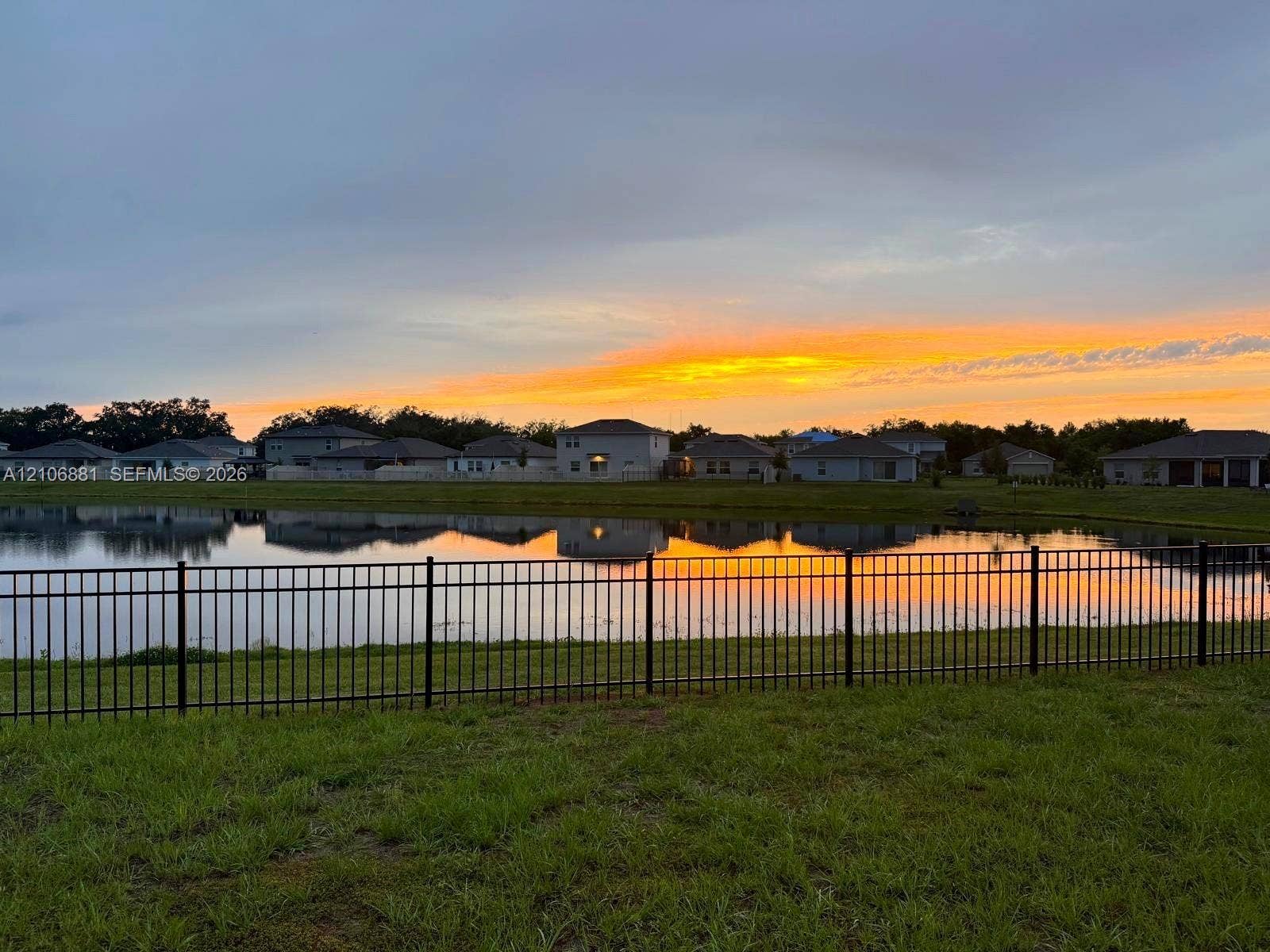 Waterfront lot featuring a black metal perimeter fence and a grass lawn, set against a backdrop of a reflective body of water