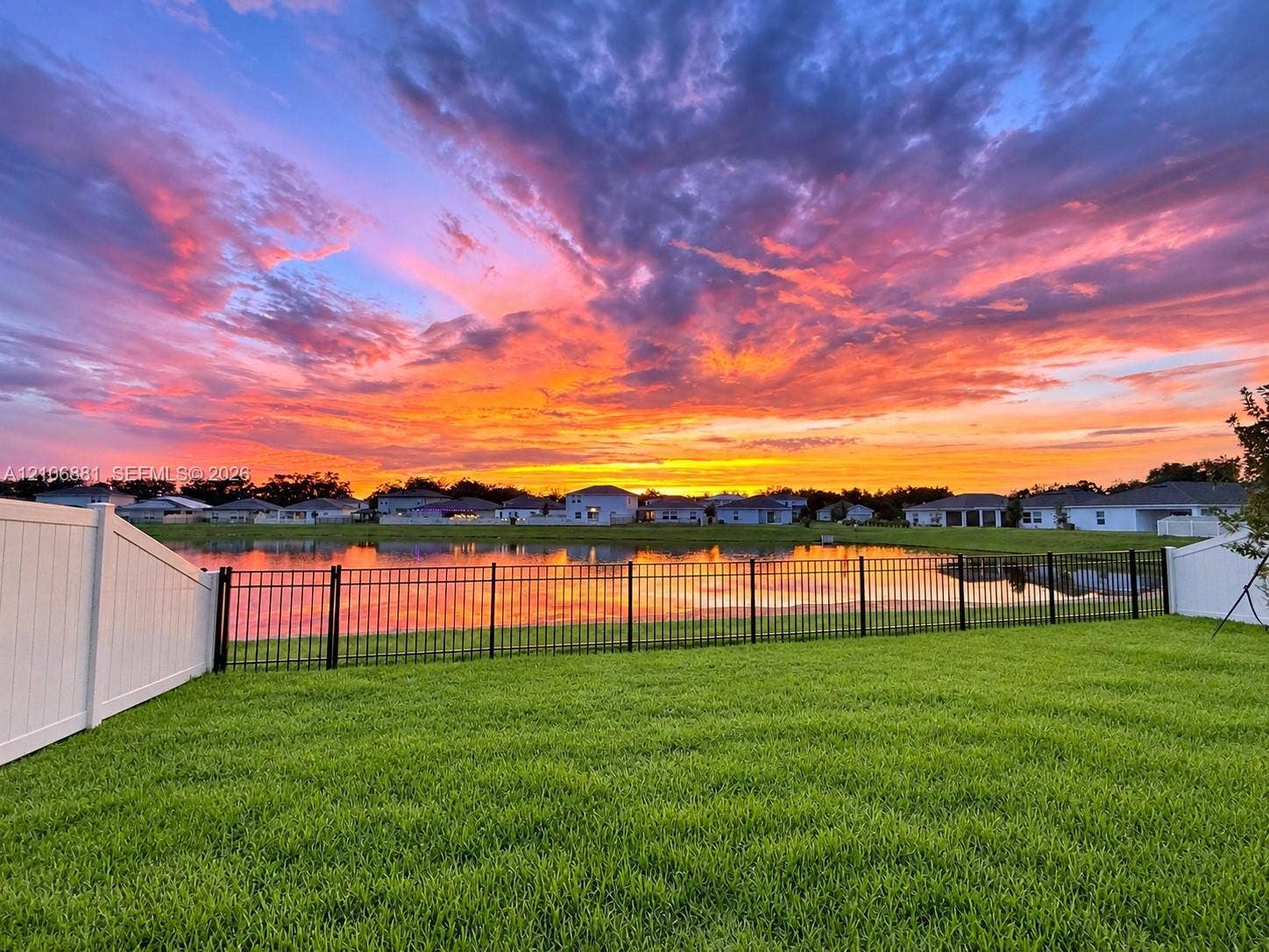 Backyard featuring a manicured lawn, water frontage, black metal perimeter fencing, and white vinyl privacy side fencing against a sunset backdrop