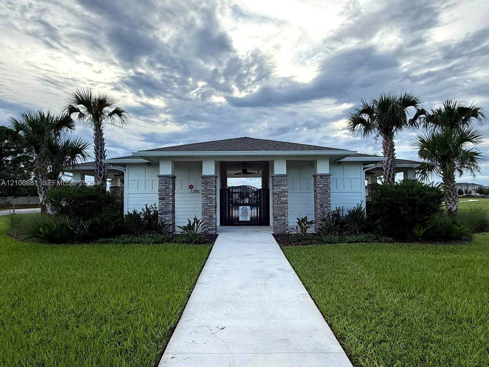 Exterior featuring stone-accented columns, white horizontal siding, a central metal gate entrance, a concrete walkway, and mature palm trees