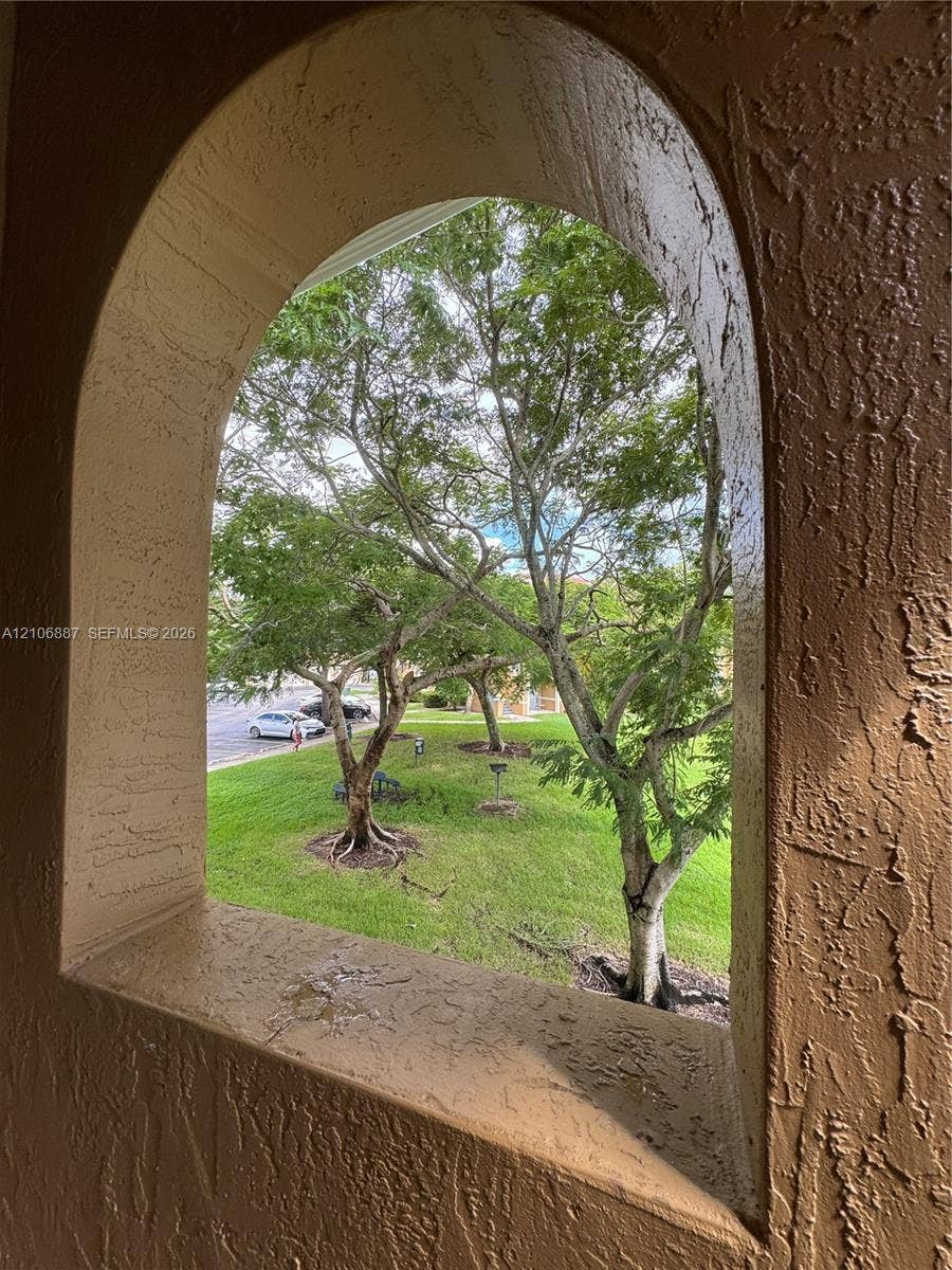 7950 N Nob Hill Road, #202, Tamarac, Tamarac, FL 33321 Arched architectural window opening with a textured stucco finish, framing a landscape featuring mature trees and a grass lawn