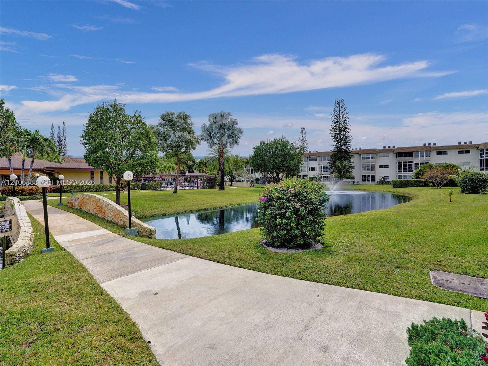3506 NW 49th Avenue, #508, Lauderdale Lakes, FL 33319 Concrete walking path featuring a stone-faced arched bridge, manicured lawn, water fountain, and mature landscaping