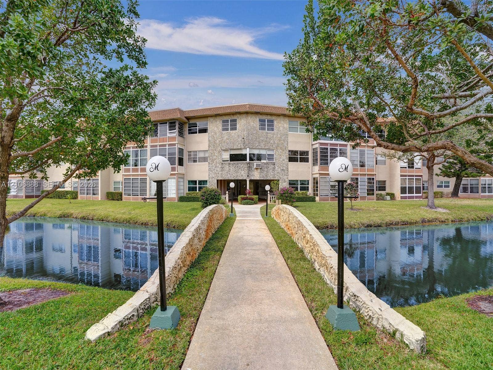 3506 NW 49th Avenue, #508, Lauderdale Lakes, FL 33319 Stone-clad bridge featuring globe-style post lighting, paved walkway, manicured lawn, and water feature