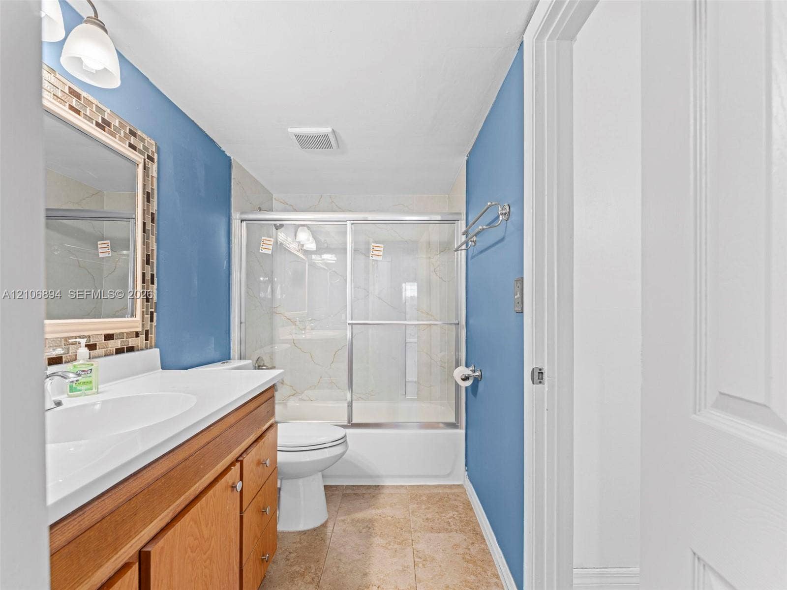 Vanity featuring a white countertop with an integrated basin, wood-finish cabinetry, a mirror with mosaic tile detailing, and a shower-tub combination with sliding glass doors and tiled wall surrounds