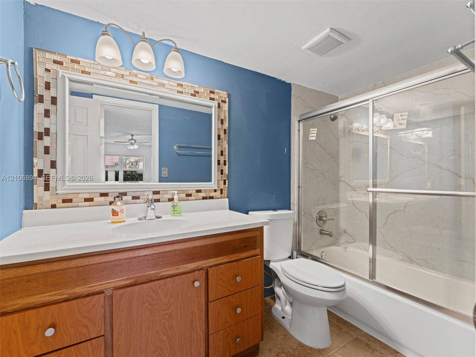 Bathroom featuring a wood-finish vanity with a white countertop, large framed mirror with mosaic tile detailing, three-light fixture, and a shower-tub combination with sliding glass doors and tiled surround